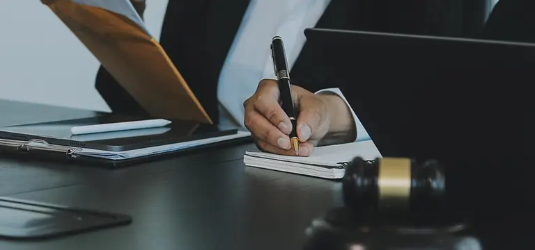 Professional in suit writing on notepad, laptop and legal documents on desk.