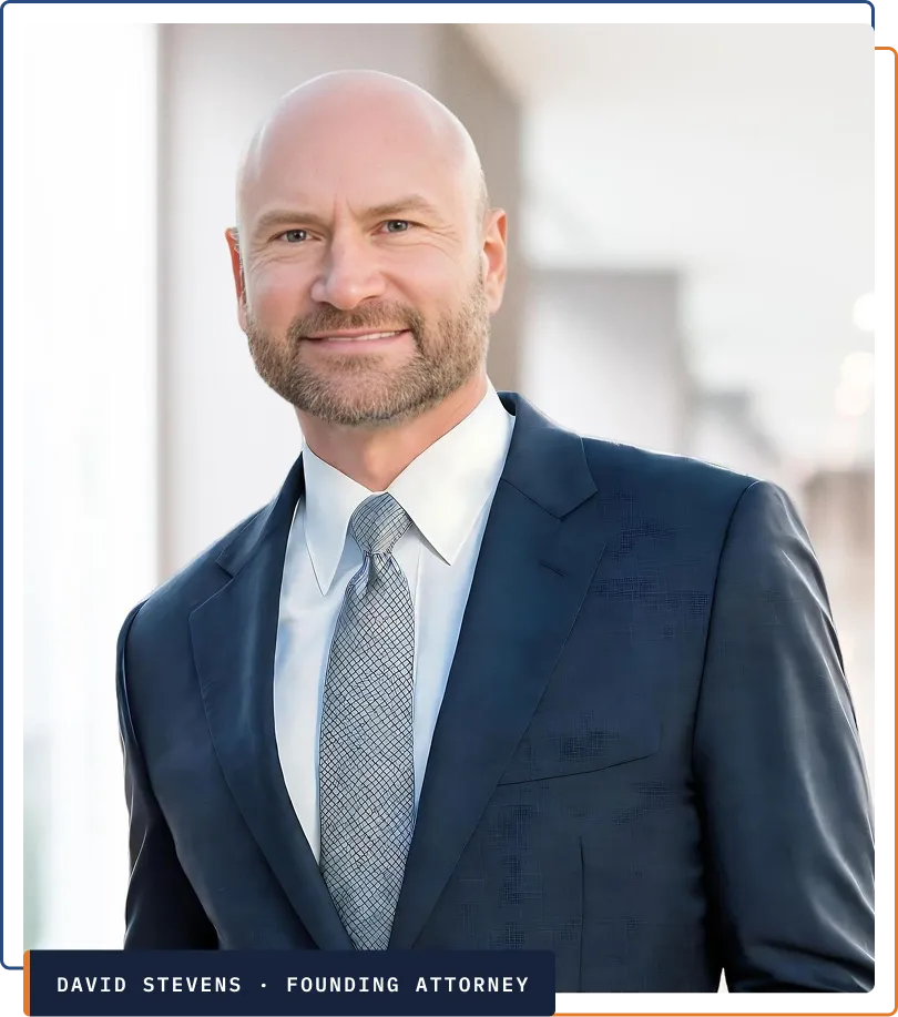 Professional portrait of a smiling bald man with a beard in a navy suit and patterned tie.