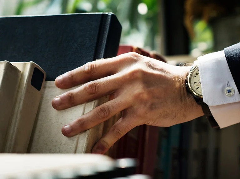 Close-up of a hand in a suit sleeve reaching for a beige book on a shelf.