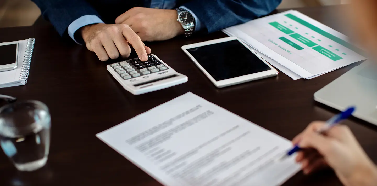 Person using a calculator on a dark wooden desk with documents, tablet, and pen.