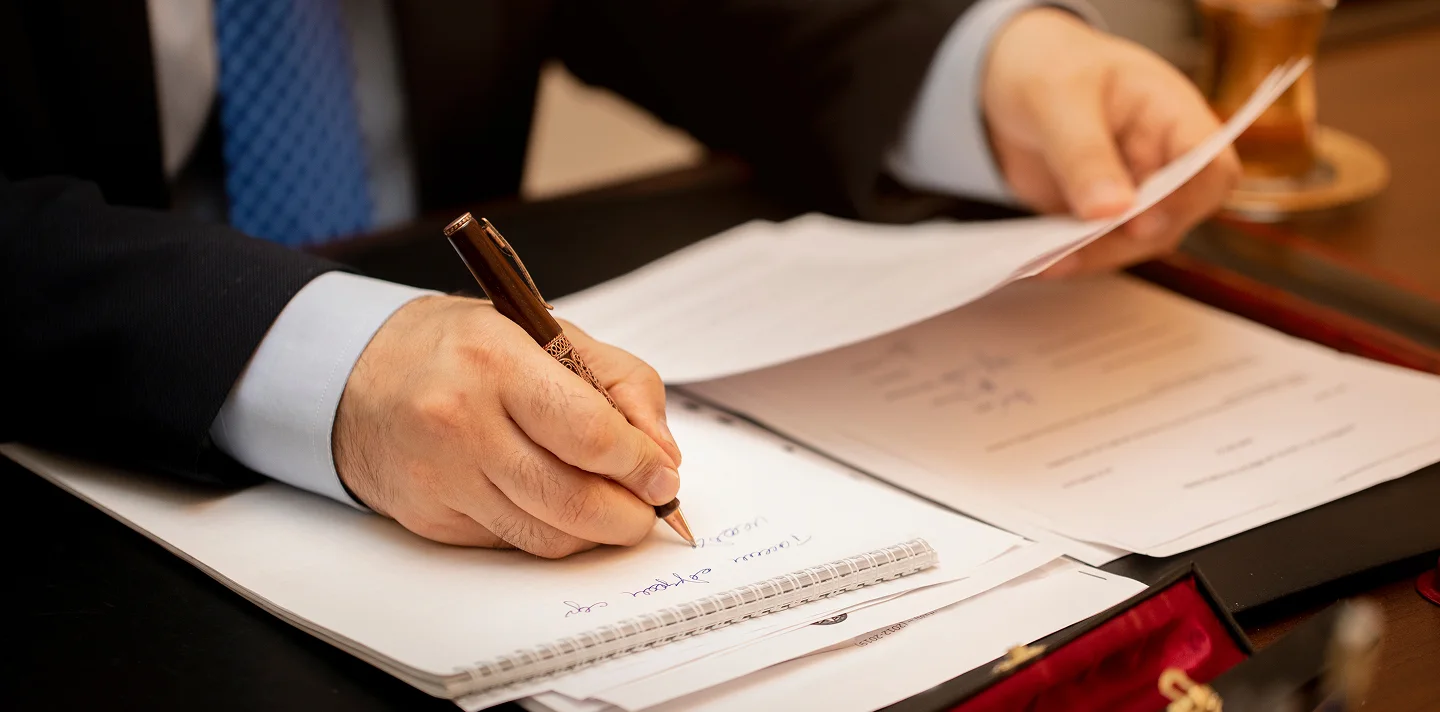 Close-up of a person in a suit writing on a notepad with documents on a desk.