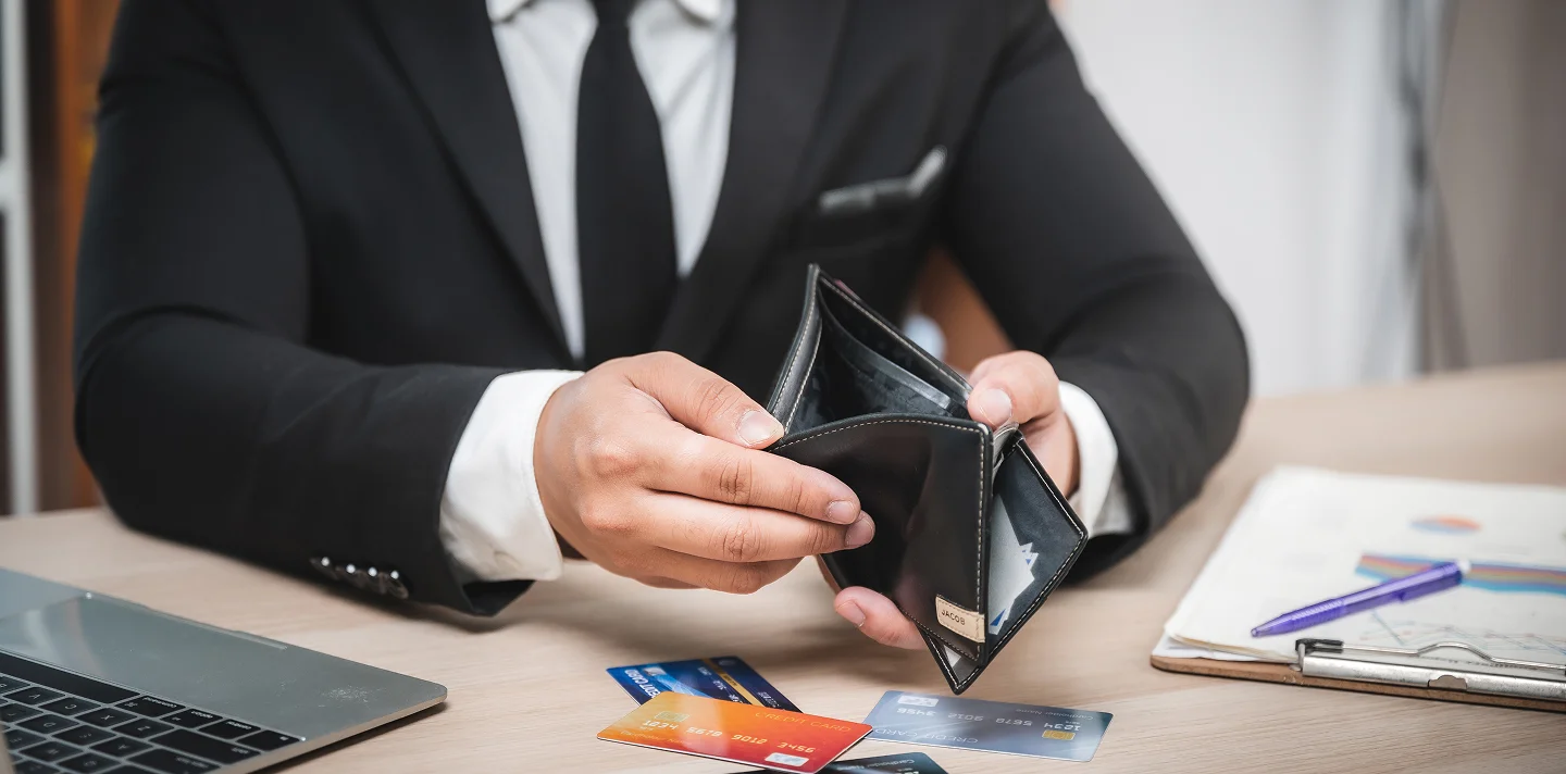 Man in black suit holding empty black leather wallet at desk with credit cards, laptop, and clipboard.