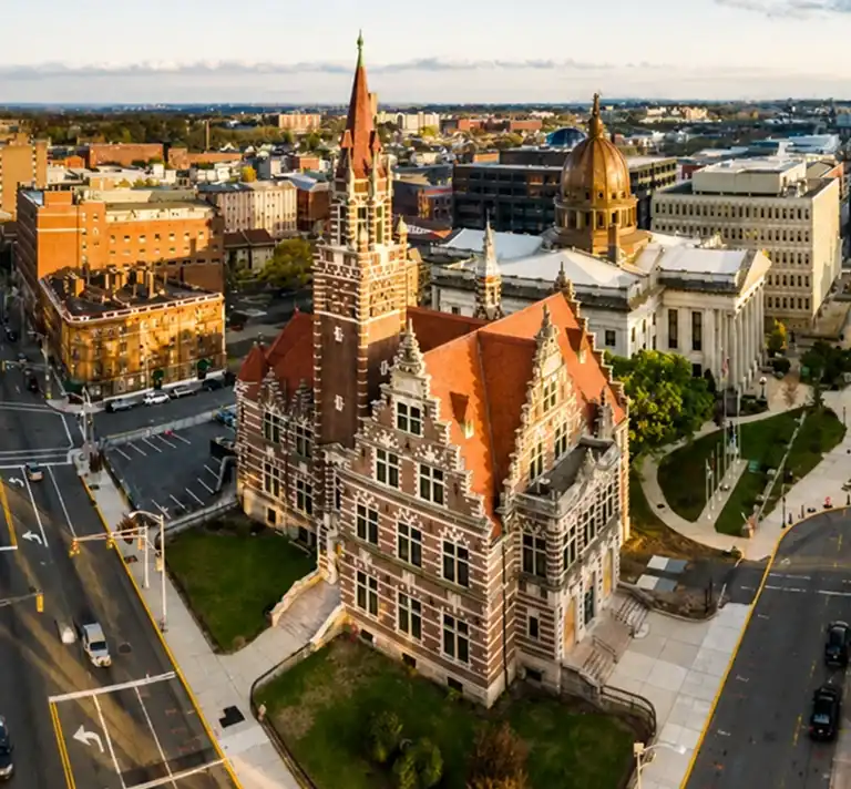 Aerial view of a historic red-brick building with a pointed tower and adjacent domed government building in an urban setting.