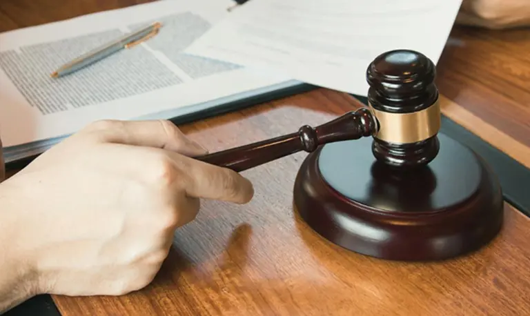 Hand holding a wooden judge's gavel above a matching sound block on a wooden desk.