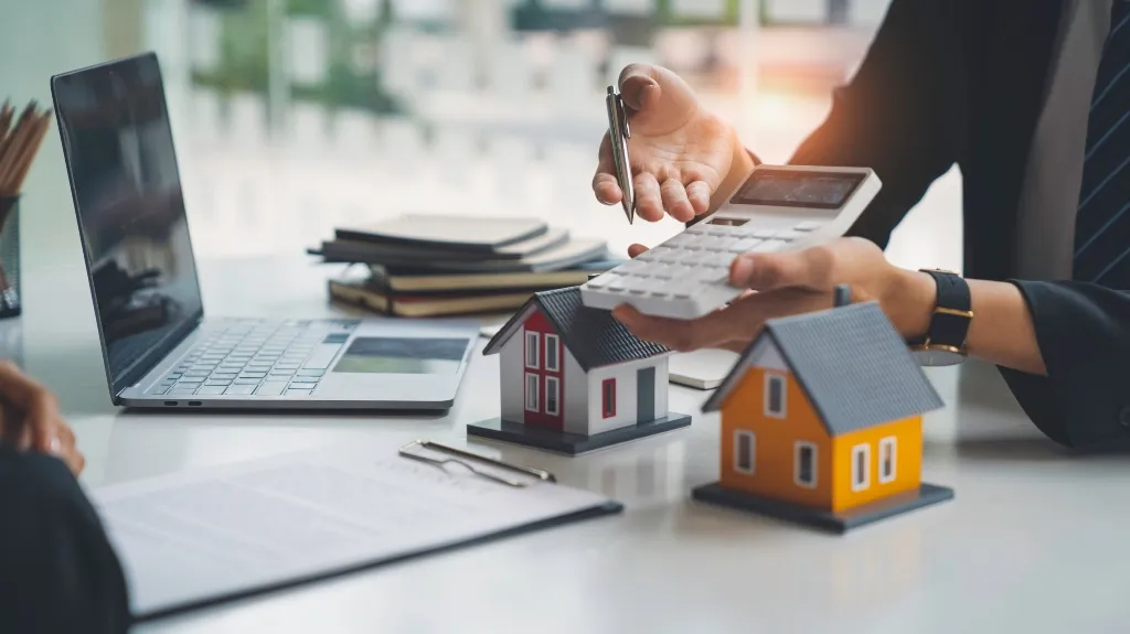 Two miniature house models on a desk with a laptop, documents, and a person holding a calculator.