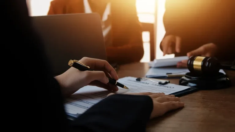 Person signing a contract document at a wooden desk with a gavel and laptop.