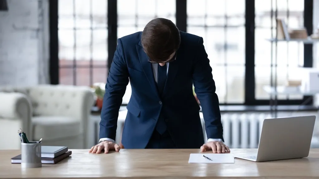 Businessman in navy suit leaning on desk with laptop and documents in modern office.