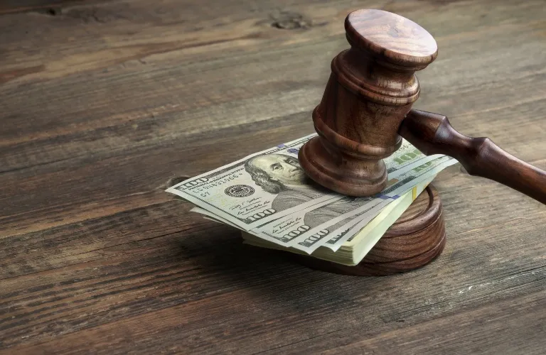 Wooden judge's gavel resting on a stack of US one hundred dollar bills on a wooden surface.