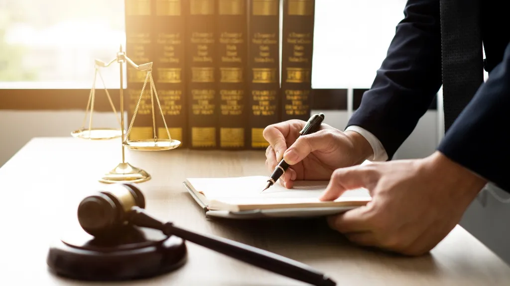 Person in formal attire writing on a document at a desk with legal books, scales of justice, and a gavel.